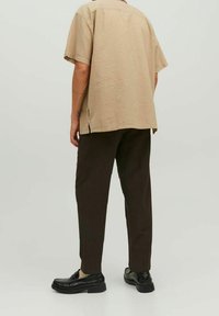 Uomo che indossa una camicia beige a maniche corte con texture, pantaloni scuri a sigaretta marroni e mocassini in pelle nera, mentre si trova in piedi contro uno sfondo neutro.