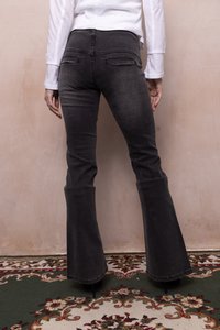 Person med højtaljede sorte flared jeans og en hvid langærmet top, stående på et mønstret tæppe mod en beige væg.