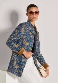 Vrouw in een blauw en beige bloemenpatroon jas, witte broek, bruine zonnebril, met een kleine beige handtas voor een effen achtergrond.