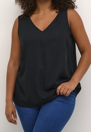 Woman with curly hair wearing a sleeveless black V-neck top and blue jeans, standing against a plain light background.