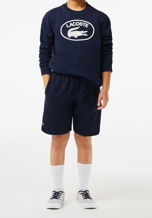 Sudadera azul marino con el logo blanco de Lacoste, combinada con pantalones cortos a juego. Mezcla de algodón, cuello redondo, banda elástica en la cintura y calcetines blancos.