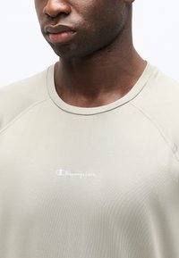 Κλείνει στο πρόσωπο ενός άνδρα που φοράει απαλό μπεζ πουλόβερ Champion με λευκό λογότυπο στο κέντρο του στήθους.