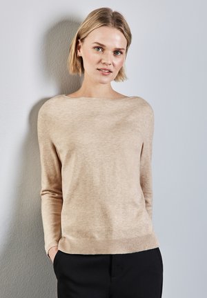 Beige Pullover mit langen Ärmeln und weitem Ausschnitt, weicher Textur, gerippten Bündchen und Saum. Das Modell posiert mit den Händen in den Taschen vor einem neutralen Hintergrund.