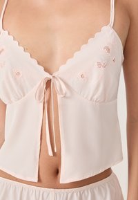 Top senza maniche di un leggero rosa con bordi scallop, ricami floreali e chiusura anteriore a cravatta. Tessuto morbido, vestibilità comoda.