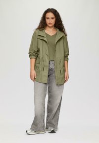 Olijfgroene jas met een capuchon, twee voorzakken en een verstelbare taille, gecombineerd met een grijze V-hals top en grijze wijde jeans. Zilveren sneakers.