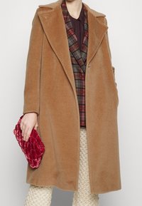 Camelkleurige wollen jas met een brede kraag en een ontspannen pasvorm. Het model houdt een rode fluwelen clutch vast, over een geruite blazer. Gelaagde broek zichtbaar.