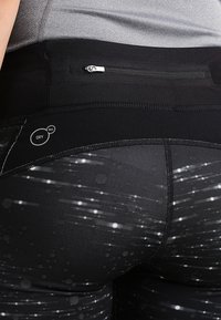 Mustat urheilulegginsit, joissa on teksturoitu pinta, vetoketjullinen tasku, heijastavat kuviot ja "DRY CELL" -logo kosteuden hallintaan.