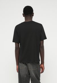 Άνδρας με κοντά σγουρά μαλλιά, φορώντας μαύρο κοντομάνικο μπλουζάκι και σκούρο γκρι τζιν, στέκεται στραμμένος μακριά από την κάμερα.