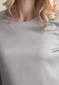 Vaalea nainen, jolla on laineikkaat hiukset, pukeutuneena yksinkertaiseen vaaleanharmaaseen pyöreäkaulaiseen paitaan, joka on leikattu olkapäistä keskivartaloon.