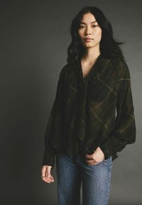 Blouse à carreaux noirs avec une coupe loose, décolleté profond et manches longues, ornée d'accents dorés et verts. Assortie avec un jean bleu.