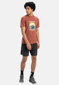 Brown short-sleeve t-shirt with a mountain graphic on the chest, paired with black shorts and gray athletic shoes.