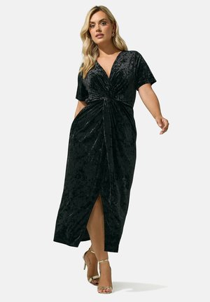 Femme aux longs cheveux blonds portant une robe portefeuille en velours noir et des sandales dorées à talons hauts, marchant devant un fond clair uni.
