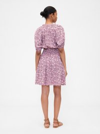 Kvinna som står med ryggen mot kameran iförd en rosa blommig knälång klänning med puffärmar och bruna sandaler.