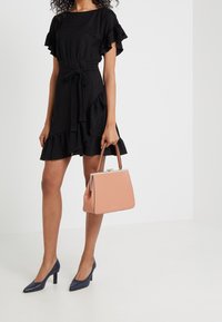 Mujer vestida con un vestido corto negro con volantes y tacones azul marino, sosteniendo un bolso estructurado de color rosa claro con asas marrones.