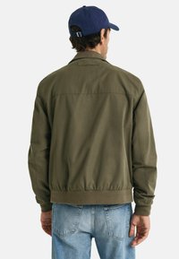 Olive green bomber jacket with elastic cuffs and waistband. Smooth fabric with a classic collar, worn with light blue jeans and a blue cap.