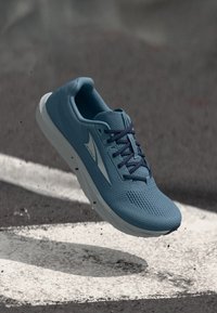 Μπλε αθλητικό παπούτσι με λευκή σόλα και σκούρα κορδόνια αιωρείται πάνω από άσφαλτο με λευκές βαμμένες γραμμές, μικρά υπολείμματα ορατά κάτω από το παπούτσι.