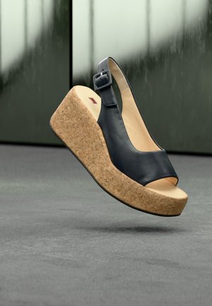 Sandalia negra de cuero con cuña y suela de plataforma de corcho y tira ajustable en el tobillo sobre suelo gris y fondo verde desenfocado.