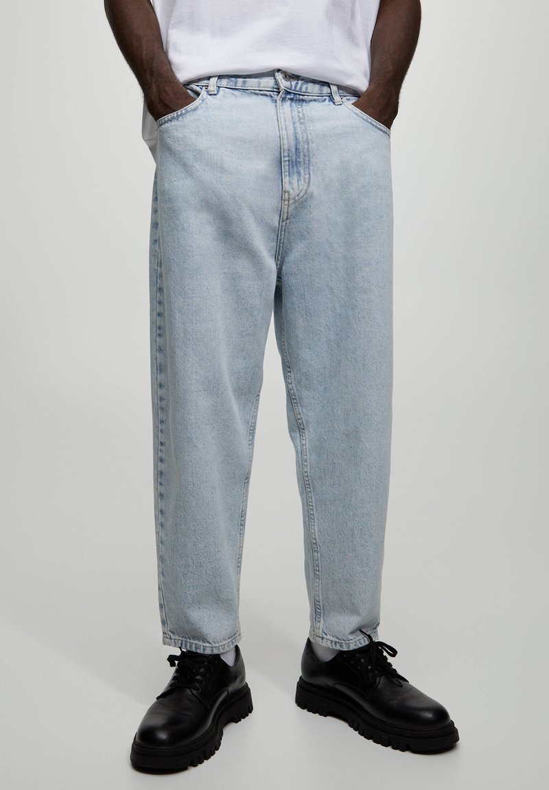 Homme portant un jean bleu clair ample, des chaussures noires épaisses à lacets, des chaussettes blanches et une chemise blanche rentrée dans le pantalon, les mains dans les poches.