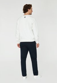 Sudadera blanca con puños y dobladillo acanalados, que presenta un pequeño logo negro en la parte posterior del cuello. Combinada con pantalones azul marino y zapatos blancos.
