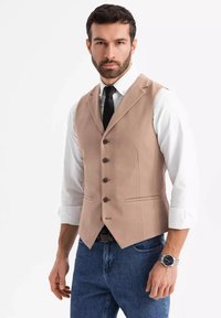 Beige strukturierte Weste mit fünf Knöpfen, zwei Taschen, getragen über einem weißen Hemd und einer schwarzen Krawatte. Kombiniert mit blauen Jeans und einer silbernen Uhr.