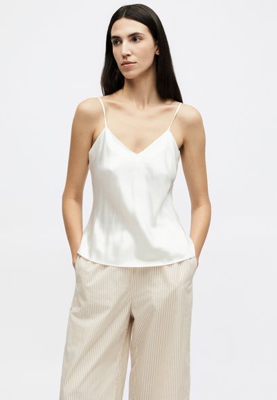 Žena s dlouhými tmavými vlasy, oblečená v bílém saténovém nátělníku a béžovo-bíle pruhovaných širokých kalhotách, ruce v kapsách, neutrální pozadí.