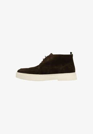 Brown suede ankle boots with a white rubber sole, featuring a lace-up design and a black pull tab at the back. Flat, smooth texture.