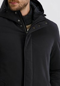 Zwarte jas met rits- en drukknoopsluiting, gewatteerde capuchon en hoge kraag. Onder de jas is een gebreide trui met textuur te zien.