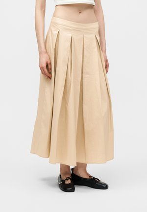 Gonna midi plissettata beige indossata con ballerine nere, mostrata su una modella dalla vita ai piedi su sfondo semplice.