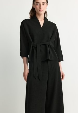 Woman wearing a black belted top with wide sleeves and matching loose black pants against a plain light background.