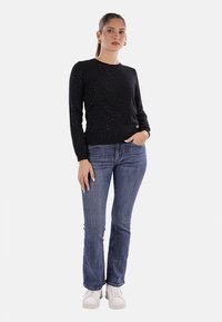 Donna in piedi che indossa un maglione a pois neri, jeans blu, sneakers bianche, con una mano in tasca su uno sfondo semplice.