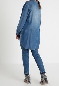 Chemise en denim bleu clair à manches longues, ourlet arrondi et fentes latérales. Associée à un jean ajusté bleu foncé et des bottines à talons blocs à motif.