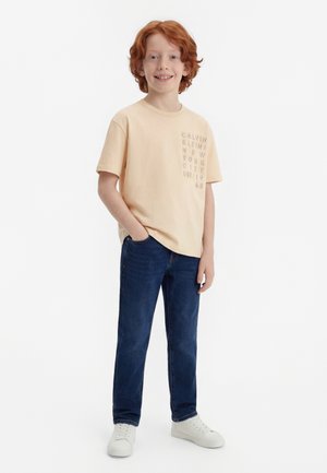 Ragazzo sorridente con capelli rossi in piedi con le mani in tasca, indossa una t-shirt beige Calvin Klein, jeans blu e sneakers bianche su sfondo bianco.