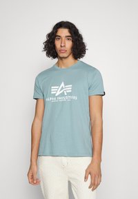 Camiseta de algodón azul claro con un logotipo gráfico blanco con formas geométricas. Cuello redondo y mangas cortas, combinada con pantalones blancos texturizados.