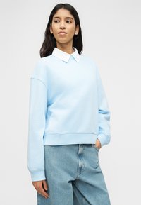 Γυναίκα που φοράει ανοιχτό μπλε φούτερ πάνω από λευκό πουκάμισο με γιακά και φαρδιά μπλε τζιν, στέκεται με το ένα χέρι στην τσέπη μπροστά σε απλό φόντο.