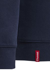 Fondo di felpa blu scuro con polsini a costine. Presenta un piccolo etichetta rossa con testo bianco, che indica il marchio "hummel". Texture morbida.