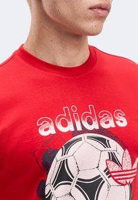 Gros plan d'une personne portant un t-shirt rouge Adidas avec un motif de ballon de football blanc et noir et le logo trèfle Adidas sur la poitrine.