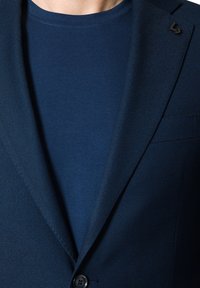 Granatowa marynarka o teksturowanej tkaninie, z pojedynczym czarnym guzikiem i klapami, noszona na dopasowanej granatowej koszulce typu crewneck.