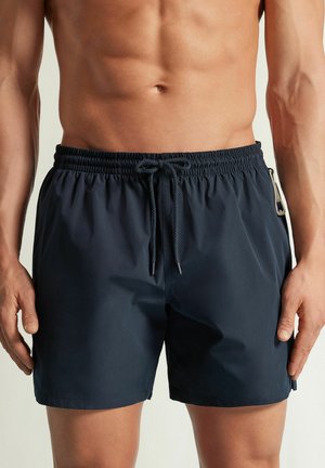 Muskularna sylwetka osoby w granatowych kąpielówkach z sznurkiem z przodu i zamykaną na zamek kieszenią boczną.