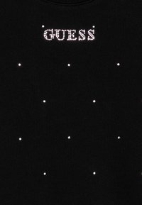 Camicia nera in tessuto con logo "GUESS" in strass luccicanti; presenta piccole pietre decorate sparse lungo la parte frontale.