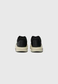 Adidași negri cu un exterior sintetic neted, accente din plasă și o talpă groasă de culoare albă-crem. Prezintă un guler căptușit și detalii de cusătură contrastante.