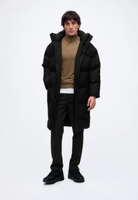 Homme debout portant un long manteau matelassé noir, un pull à col roulé marron, un pantalon noir et des chaussures noires sur un fond blanc uni.