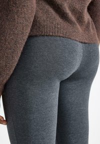 Tmavě šedé legíny vyrobené z elastické tkaniny, přiléhavý střih, hladká textura. Nahoře je částečně vidět hnědý pletený svetr.