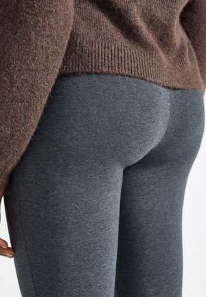 Mørkegrå leggings lavet af strækbart stof, tætsiddende design, glat tekstur. Båret med en brun strikket trøje, delvist synlig for oven.
