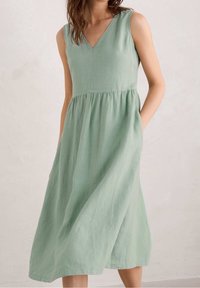 Robe sans manches en lin vert menthe avec décolleté en V et taille froncée, portée par une personne les mains dans les poches, debout contre un mur clair.