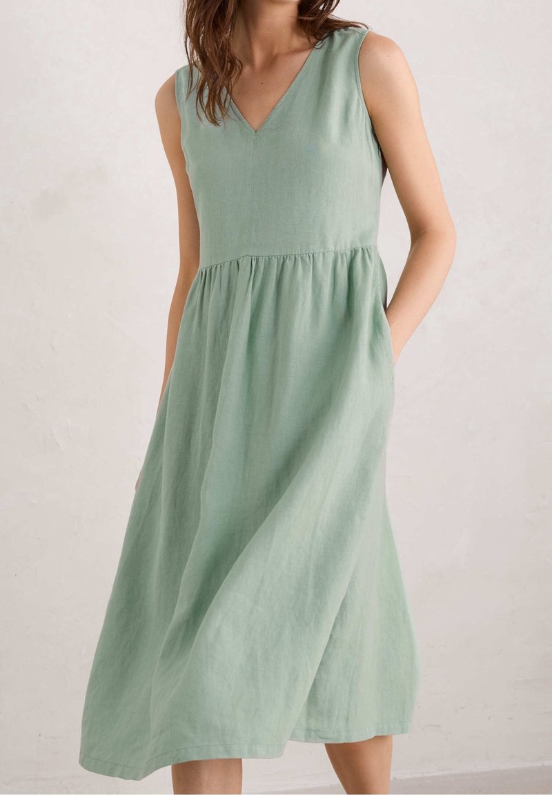 Robe sans manches en lin vert menthe avec décolleté en V et taille froncée, portée par une personne les mains dans les poches, debout contre un mur clair.