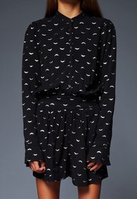 Vestido negro con un escote redondo, cierre de botones y mangas largas. Presenta una tela texturizada con un patrón de hojas blancas. La cintura está ajustada.