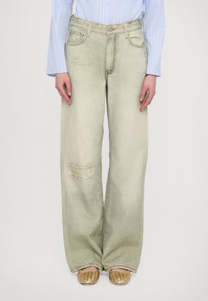 Personne portant un jean large vert clair usé, une chemise rayée bleue et des chaussures tissées beiges sans lacets, debout devant un fond uni.