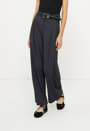 Άτομο που φοράει ψηλόμεσο σκούρο παντελόνι με φαρδύ πόδι, μαύρη ζώνη, μαύρο μαν sleeveless τοπ και μαύρα μπαλαρίνια με λευκές λουρίδες, όρθιο σε λευκό πάτωμα.