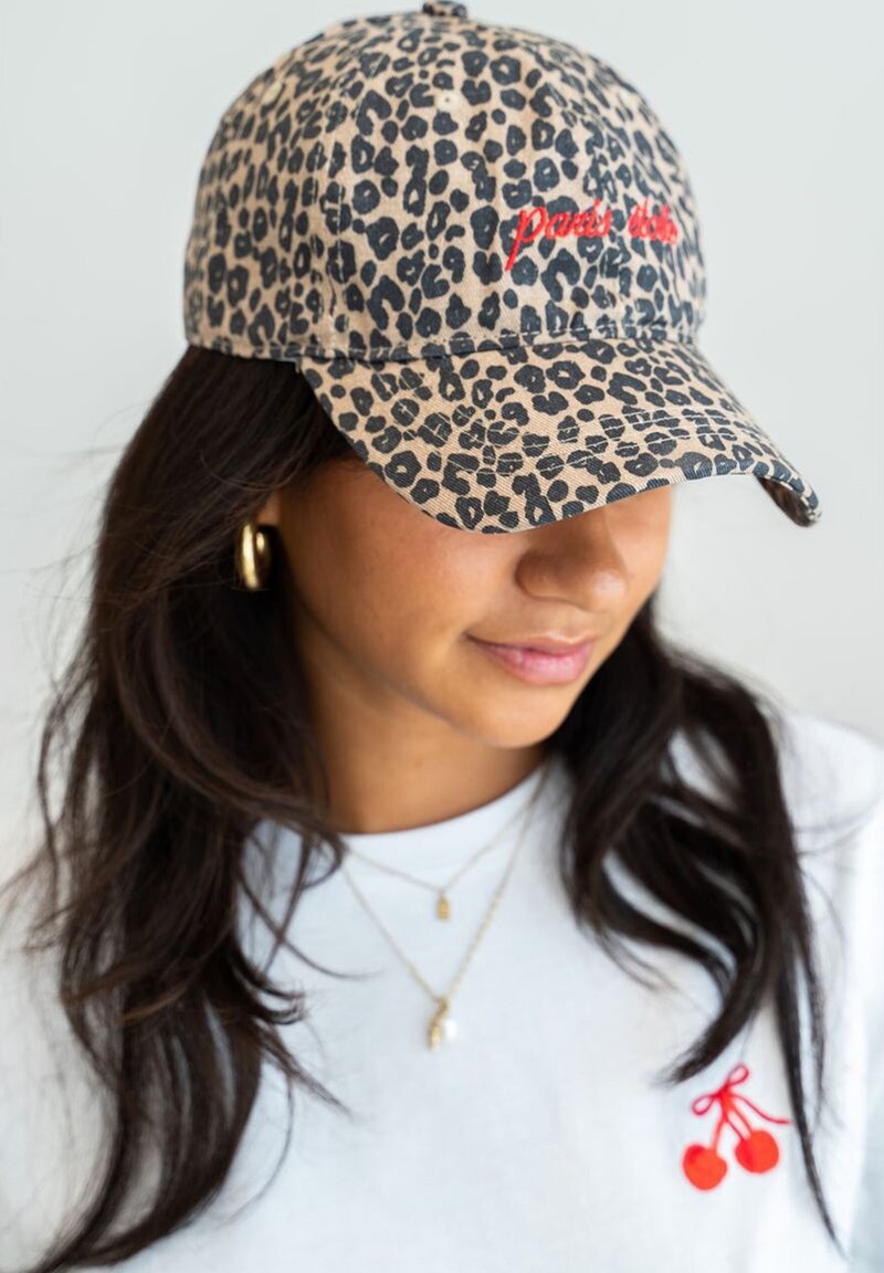 Leopard print baseball cap in tan and black, featuring red embroidered text. Soft fabric with a curved brim and adjustable strap.