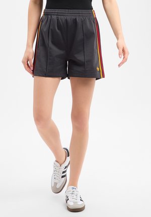 Personne portant un short de sport noir avec des bandes latérales rouges et jaunes et des baskets blanches à bandes noires, debout sur fond blanc.